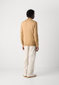 Man die met zijn rug naar de camera staat, gekleed in een beige longsleeve trui, off-white broek en witte sneakers, tegen een witte achtergrond.