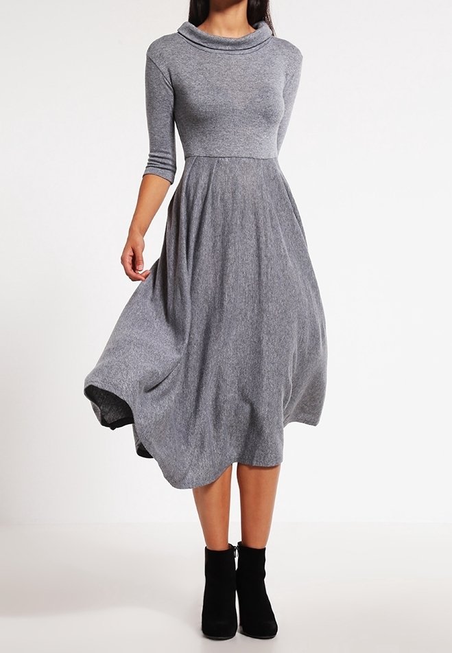 Femme portant une robe en maille grise mi-longue à manches trois-quarts et des bottines noires, debout devant un fond uni.