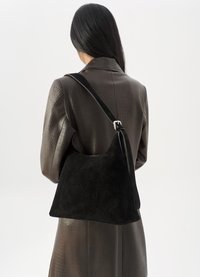 Woman with long dark hair wearing a brown textured coat carries a large black suede shoulder bag with an adjustable strap.