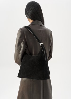 Woman with long dark hair wearing a brown textured coat carries a large black suede shoulder bag with an adjustable strap.