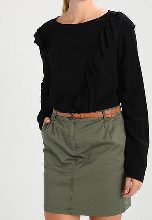 Femme portant un haut noir à manches longues avec des volants, rentré dans une jupe vert olive avec une ceinture marron, main dans la poche.