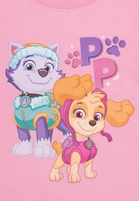 Personaggi dei cartoni animati: un lupo viola con accenti blu e un cane color sabbia in un vestito rosa, su uno sfondo rosa con impronte di zampe e lettere.