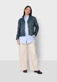 Tmavě zelená kožená bunda se zkráceným střihem, dvěma předními kapsami a zapínáním na knoflíky, spárovaná s světle modrou košilí a krémovými kalhotami s širokými nohavicemi.