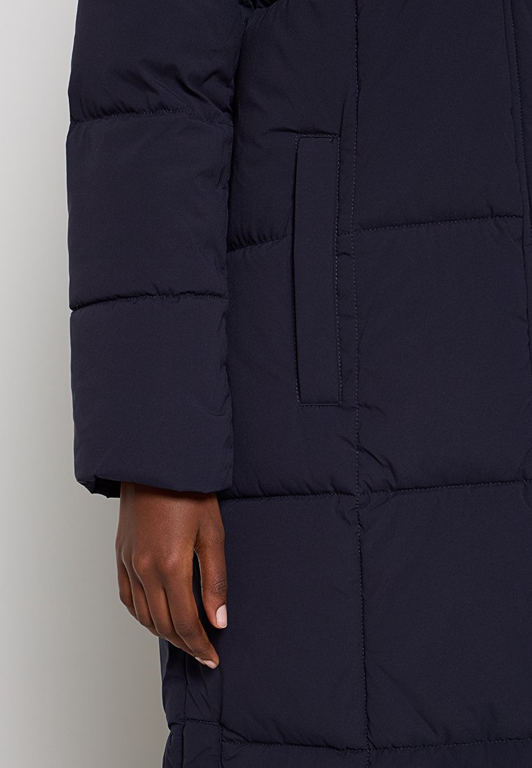 Cappotto imbottito blu navy con design trapuntato, dotato di una tasca laterale e maniche strutturate. Il materiale sembra liscio e isolante.