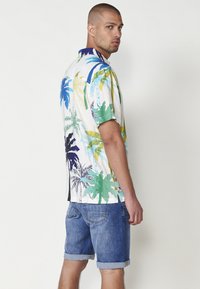 Cămașă cu mânecă scurtă și guler, având un imprimeu tropical vibrant cu palmieri în nuanțe de albastru, verde și galben pe un fundal alb; șorturi din denim.