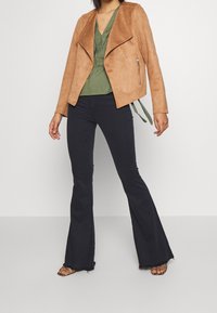 Donna che indossa una giacca in suede beige, un top avvolgente verde oliva, pantaloni neri a zampa d'elefante e sandali con le dita scoperte, in piedi su uno sfondo semplice.