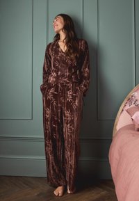 Dunkelbraunes, samtiges Damenoutfit mit einem Wickeloberteil und weit geschnittenen Hosen, die ein dezentes, helles Blattmuster aufweisen.