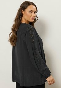 Sort bluse med lange ærmer, prydet med indviklet broderi langs ærmerne. Fremstillet af et let, flowy materiale.