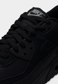 Μαύρο αθλητικό παπούτσι Nike με επάνω μέρος με υφή και λείες επικαλύψεις, μαλακή σόλα, ορατό κορδόνι και λογότυπο στη γλώσσα.