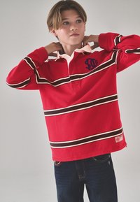 Camisa polo roja de manga larga con cuello de solapa, con rayas horizontales blancas y marrones, y un logo en el pecho izquierdo. Combinada con vaqueros azules.