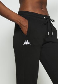 Detailní záběr osoby v černých teplácích s bílým logem na kapse, ruka ležérně zasunutá v kapse.