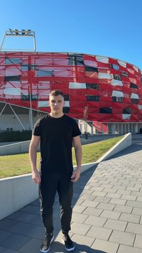 Čierne tričko a nohavice na osobe stoja na dláždenej ceste s červeným a bielym vzorovaným štadiónom v pozadí pod jasnou modrou oblohou.