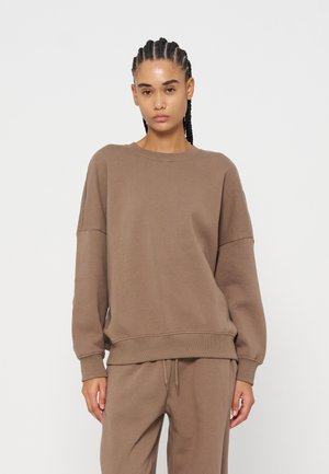 Femme aux cheveux tressés portant un sweat-shirt marron ample et un pantalon de jogging assorti sur fond blanc uni.