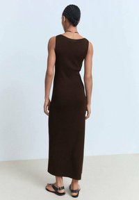 Hnědá maxi šaty bez rukávů, pletené, s kulatým výstřihem a přiléhavým střihem. Modelka má na sobě černé sandály. Pozadí je světle modré.