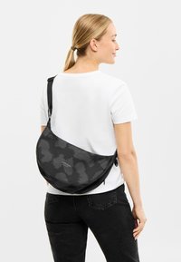 Geantă de umăr cu model negru, de formă curbată, având un logo, bretea ajustabilă și materiale netede din nylon, purtată pe umăr.