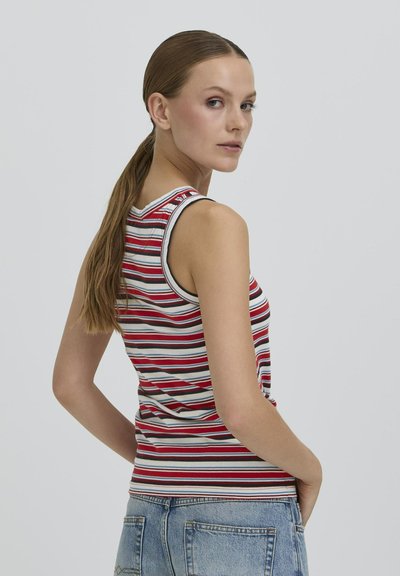 Femme avec une longue queue de cheval portant un haut sans manches rayé rouge, blanc et bleu et un jean en denim bleu clair, regardant par-dessus son épaule.