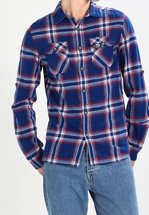 Muž oblečený v modré, červené a bílé kárované košili s knoflíkovými kapsami na hrudi a vyhrnutými rukávy, spárovaný s modrými džínami.