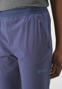 Lichte paarse wandelbroek met een elastische tailleband en een ritszak aan de zijkant. Bevat een logo op de dij en heeft een gladde textuur.