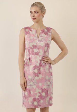 Woman wearing sleeveless, knee-length dress with pink and white large floral pattern, standing against plain beige background.