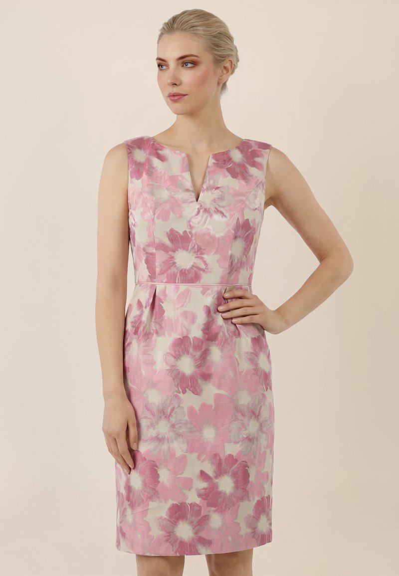 Frau in einem ärmellosen, knielangen Kleid mit großem pink-weißem Blumenmuster, stehend vor einem einfarbig beigen Hintergrund.