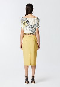 Žuta suknja do koljena s prorezom straga, uparena s cvjetnom bluzom s otvorenim ramenima koja ima crne i žute uzorke. Crne štikle.