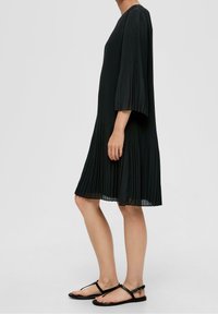 Svart plisserad klänning med trekvartslånga klockärmar, knälång, tillverkad av lättviktig stof; tillsammans med svarta platta sandaler.