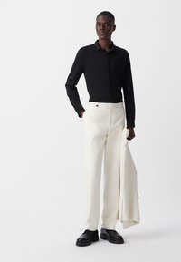 Man in een zwarte lange mouw shirt en witte broek, met een bijpassende witte jas in de hand, staande tegen een effen witte achtergrond.