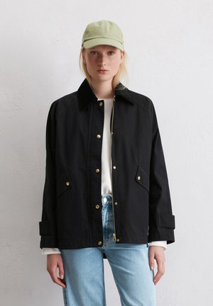 Black jacket with gold hardware, buttoned pockets, and a collar, paired with a light green cap. Model wears light blue jeans and a white shirt.