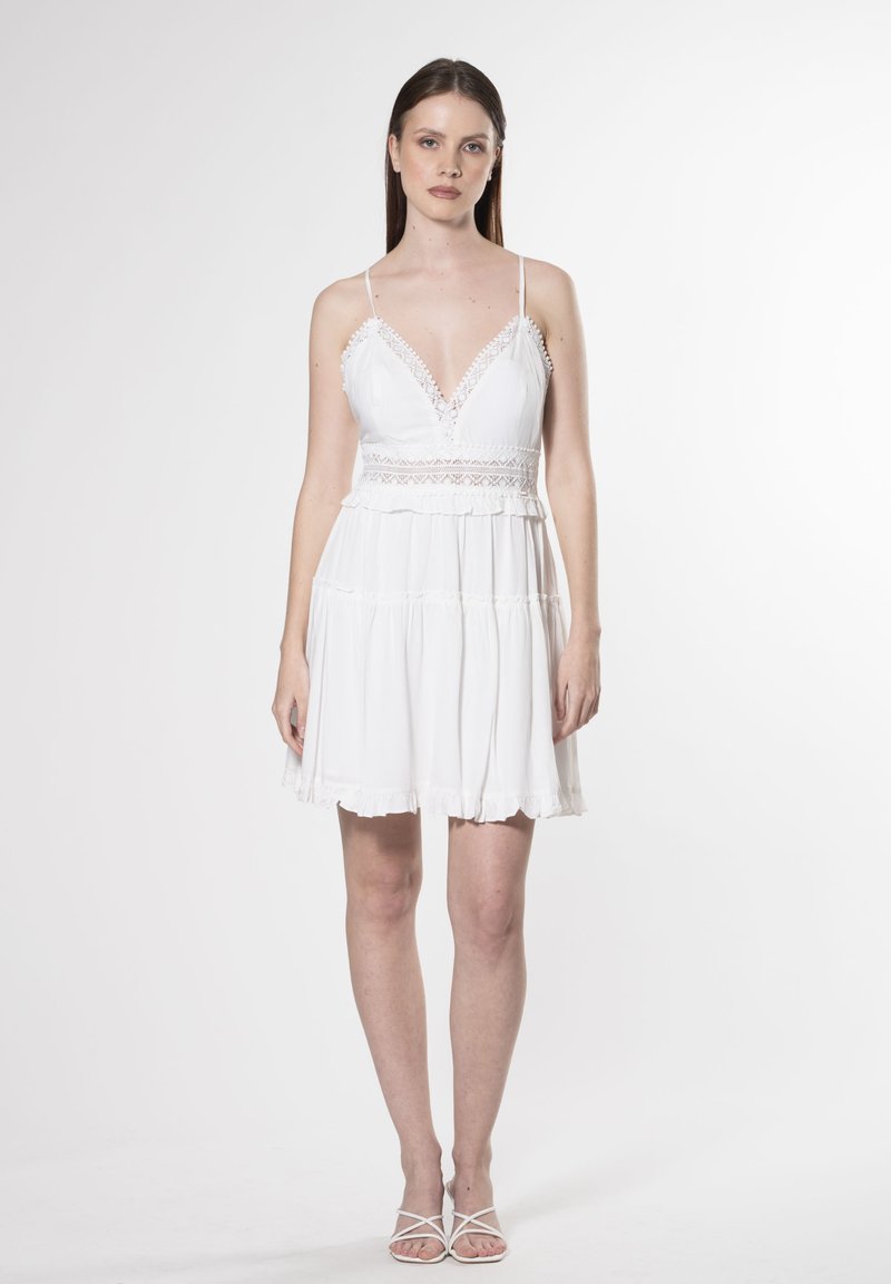 Vestido mini blanco con escote en V, borde de encaje, tirantes finos ajustables y dobladillo con volantes en capas. Tejido ligero con una textura suave.