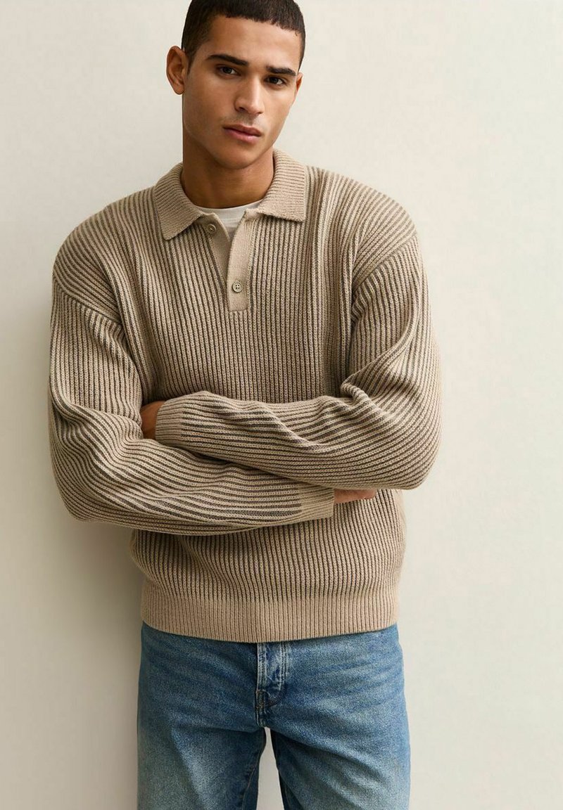 Młody mężczyzna z krótkimi włosami, ubrany w beżowy, prążkowany sweter na białej koszuli i niebieskich dżinsach, stojący z założonymi rękami na tle jednolitego tła.