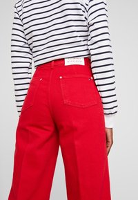 Persona che indossa pantaloni rossi a gamba larga e a vita alta e una camicia bianca a maniche lunghe con strisce orizzontali nere, vista da dietro.