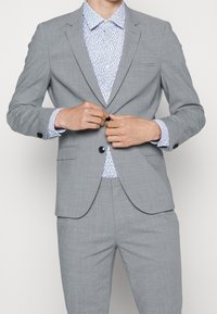 Traje gris claro con un corte entallado. La chaqueta tiene un cierre de un solo botón, solapas con muesca y una camisa texturizada con patrón azul y blanco debajo.