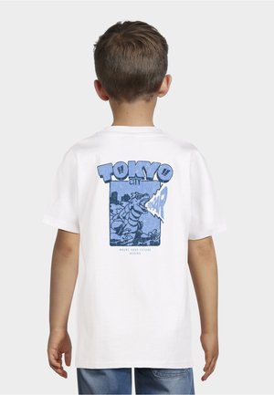 Dreng iført hvid T-shirt med blå grafik af en brølende dinosaurus og teksten "Tokyo City" på ryggen mod en ensfarvet baggrund.