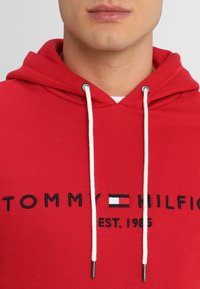Sudadera roja con capucha ajustable, que presenta texto negro bordado "TOMMY HILFIGER EST. 1985" y acento de logo. Tejido de algodón suave.