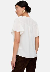White short-sleeve blouse with ruffled accents, soft texture, a relaxed fit, and a rounded back hem. Gold earrings are also visible.