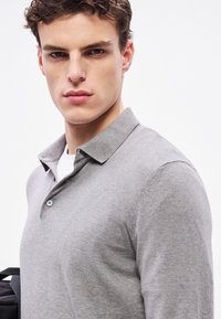 Tricou polo gri deschis, din material moale, cu guler și guler cu nasturi. Modelul are părul scurt și ține o geantă neagră.