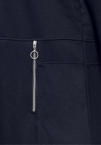 Le tissu du sweat navy présente une poche latérale avec une fermeture éclair argentée et un tirette circulaire, mettant en avant un design épuré et une texture lisse.
