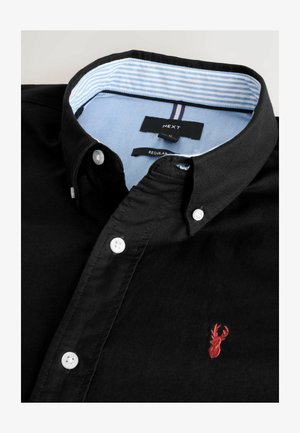 Fekete pamut ing fehér gombokkal, galléros kialakítással, hímzett piros szarvaslogóval a mellkason és világoskék csíkozott belső gallérral.