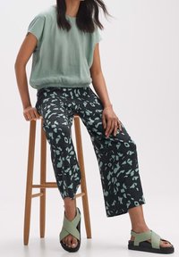 Kvinna som sitter på en träpall, iklädd en mintgrön överdel, byxor med svart och grön mönster, samt ljusgröna sandaler med korsremmar på vit bakgrund.