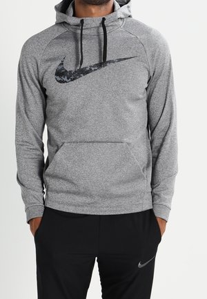 Uomo che indossa una felpa con cappuccio grigia Nike con logo swoosh mimetico digitale e pantaloni neri Nike con logo swoosh, mani nelle tasche.