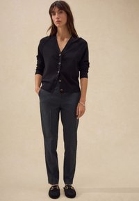 Cardigan nero con scollo a V e chiusura con bottoni, abbinato a pantaloni scuri dalla trama texturizzata. Il modello indossa scarpe scure con dettagli in oro.