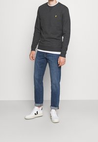 Felpa grigia con logo giallo, abbinata a jeans blu arrotolati e sneakers bianche con dettagli neri. Design pulito e casual.