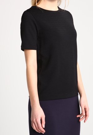 Žena v přiléhavém černém texturovaném tričku s krátkým rukávem a tmavě modré sukni stojí proti jednoduchému světlému pozadí.