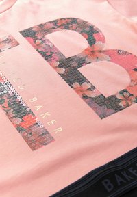 Lyserød bomulds T-shirt med et stort blomsterprintet grafik "B" og guldtekst, med en tekstureret paliet-detalje.