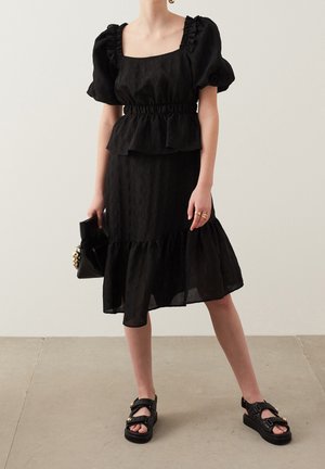 Kobieta ubrana w czarną, teksturowaną bluzkę z bufkami i dopasowaną spódnicę midi, trzymająca czarną kopertówkę i nosząca czarne sandały ze złotymi detalami.