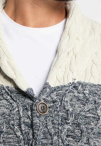 Cárdigan de punto trenzado que presenta una sección superior color crema y una sección inferior de un gris oscuro, con un gran botón y detalle de cuello acanalado.