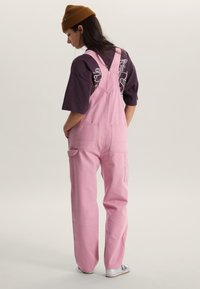 Roze denim tuinbroek met verstelbare bandjes, twee grote zijzakken en een losse pasvorm. Draagt over een donker T-shirt met print en witte sneakers.