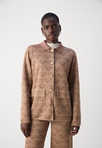Musterjacke in Hellbraun, mit einem Kragen, goldenen Knöpfen und zwei vorderen Taschen. Getragen über einem strukturierten Oberteil mit einem rüschierten Halsausschnitt.
