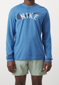 Muž v modrém tričku s dlouhým rukávem Nike a světle zelených šortkách Nike, stojící s uvolněnýma rukama podél těla na jednobarevném bílém pozadí.