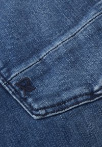 Donkerblauwe denimstof met een subtiele textuur. Bevat een klein geborduurd 'R' logo en zichtbare stiksels langs de randen.